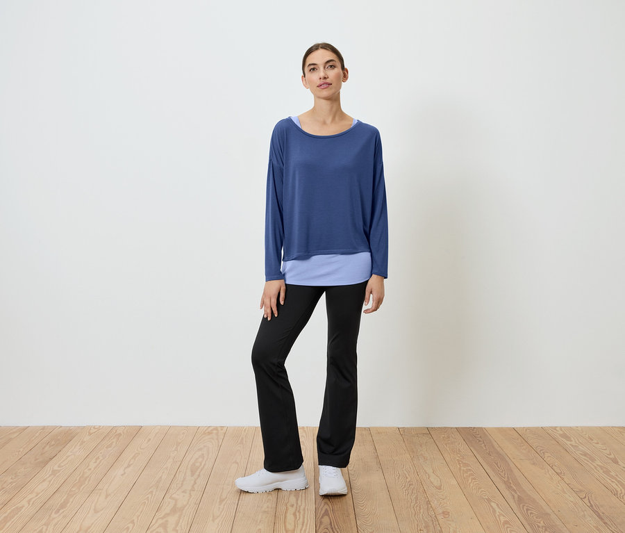 Une femme se tient sur un plancher en bois, portant un haut bleu et un pantalon noir.