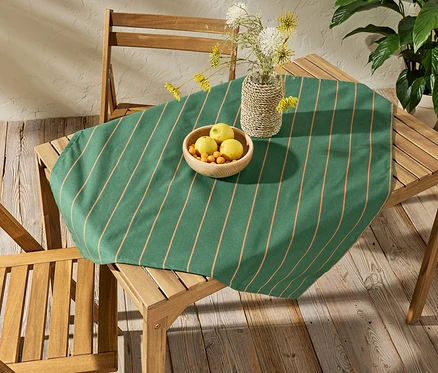 Sur une table en bois se trouve une nappe lavable d'environ 90 x 90 cm à rayures. Sur la nappe, il y a un bol de fruits et un vase avec des fleurs.