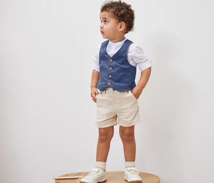 Un petit garçon debout sur un tabouret en bois porte un gilet pour enfant, une chemise, un short et des chaussures.