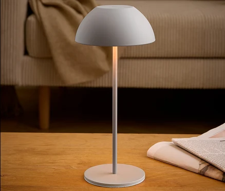 Une lampe grise est posée sur une table en bois à côté d'un journal. Un canapé beige est visible à l'arrière-plan.