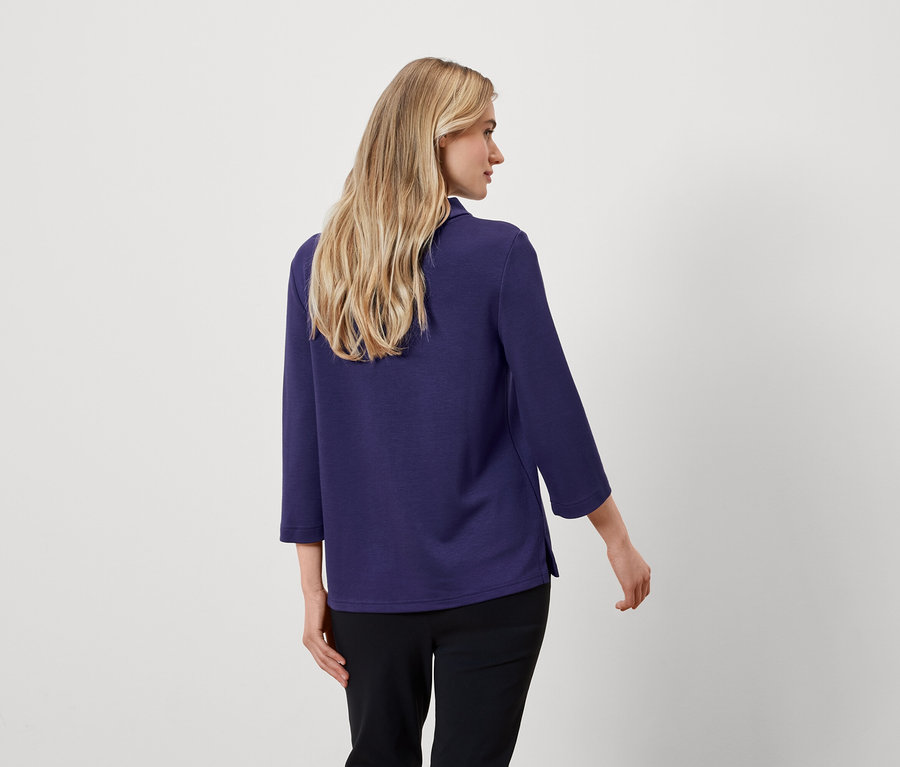 De dos, une femme blonde porte un chemisier bleu à manches trois-quarts et un pantalon noir.