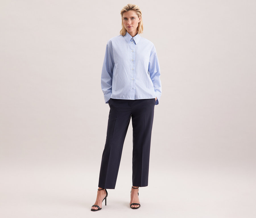 Femme portant un chemisier oversize Seidensticker à rayures bleues, un pantalon foncé et des sandales noires à talons.
