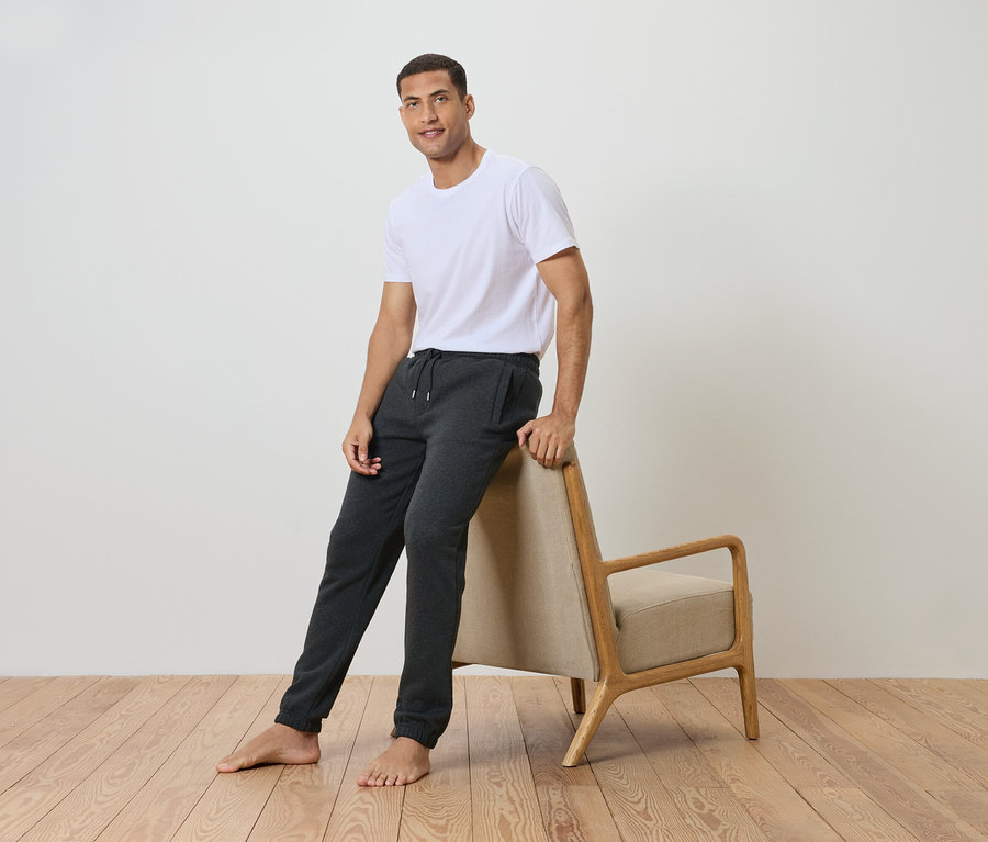 Un homme appuyé contre une chaise, portant un t-shirt blanc et un pantalon de jogging gris.