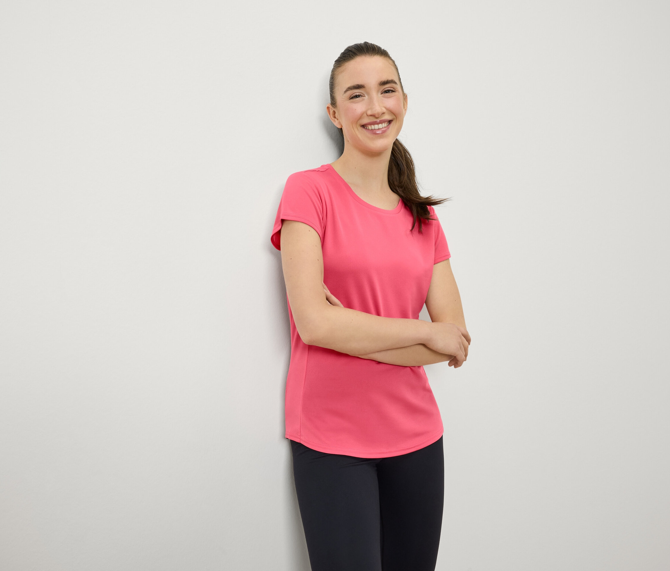 Une femme souriante aux cheveux bruns coiffés en queue de cheval se tient debout, les bras croisés sur la poitrine. Elle porte un t-shirt rose et un legging bleu marine.