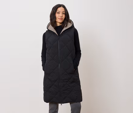 Une femme porte un long gilet noir matelassé à capuche.