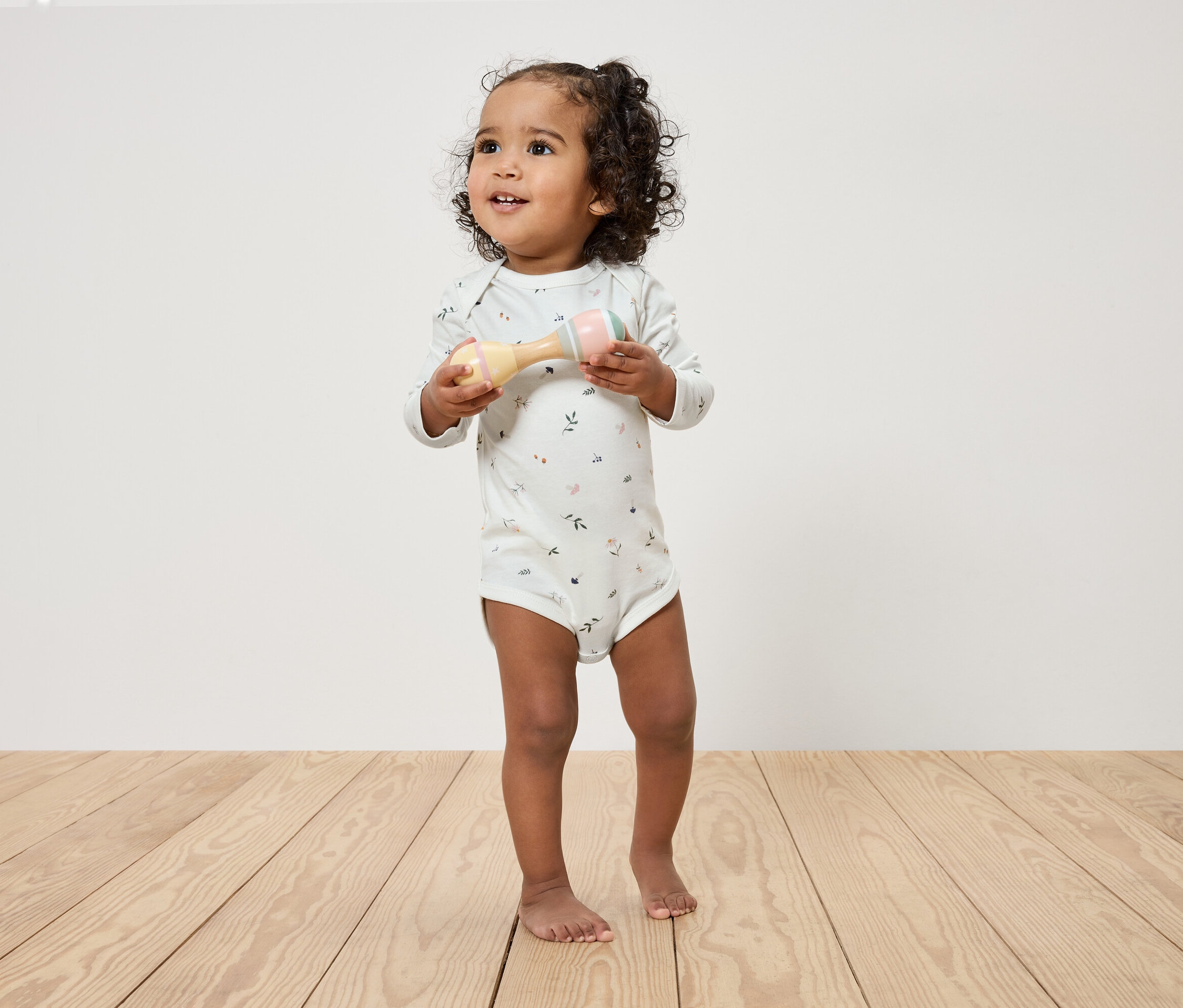 Un enfant aux cheveux bouclés tient un hochet dans ses mains et se tient sur un plancher en bois devant un mur blanc.