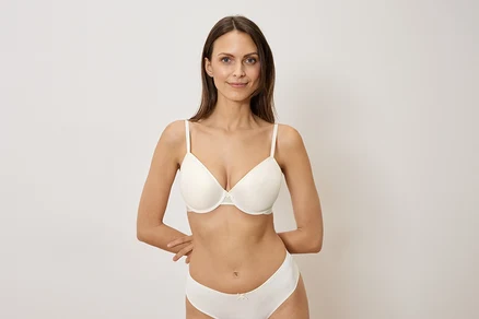 Femme aux longs cheveux bruns portant un soutien-gorge et des sous-vêtements blancs posant devant un fond blanc.