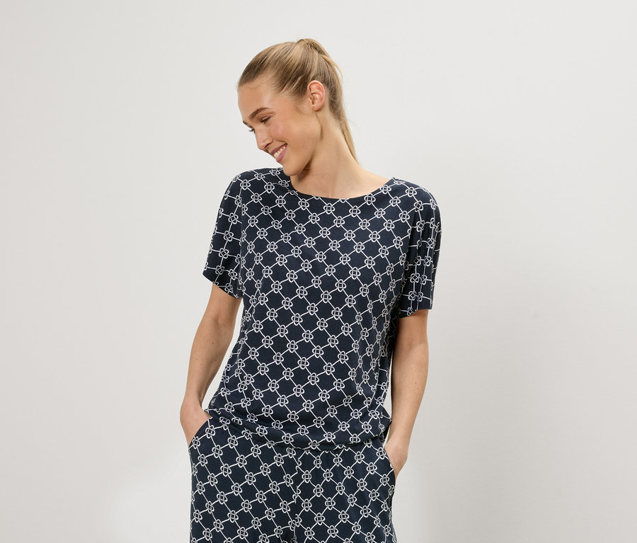Une femme blonde porte un haut bleu à manches courtes avec un motif géométrique blanc et un pantalon assorti.