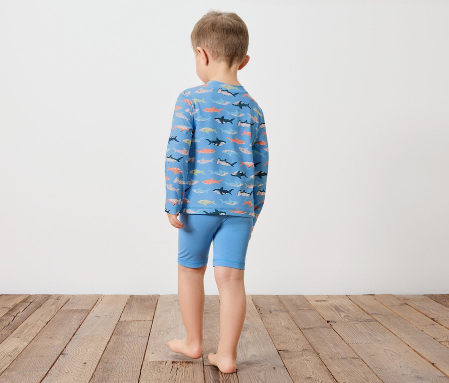 Un enfant se tient dos à la caméra, il porte un haut bleu à manches longues avec un motif de requins et un short bleu.
