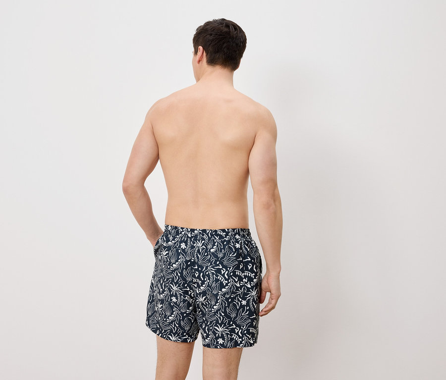 Homme vu de dos, portant un short de bain avec un motif blanc sur fond bleu, la main dans la poche.