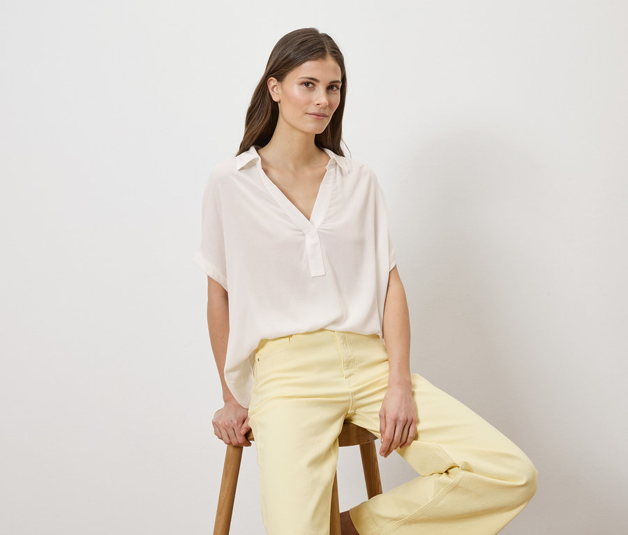 Une femme est assise sur un tabouret, portant un chemisier blanc et un pantalon jaune.