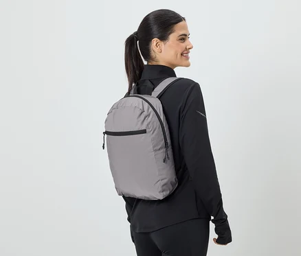 Une femme avec une queue de cheval porte un sac à dos gris et des vêtements de sport noirs, vue de côté.
