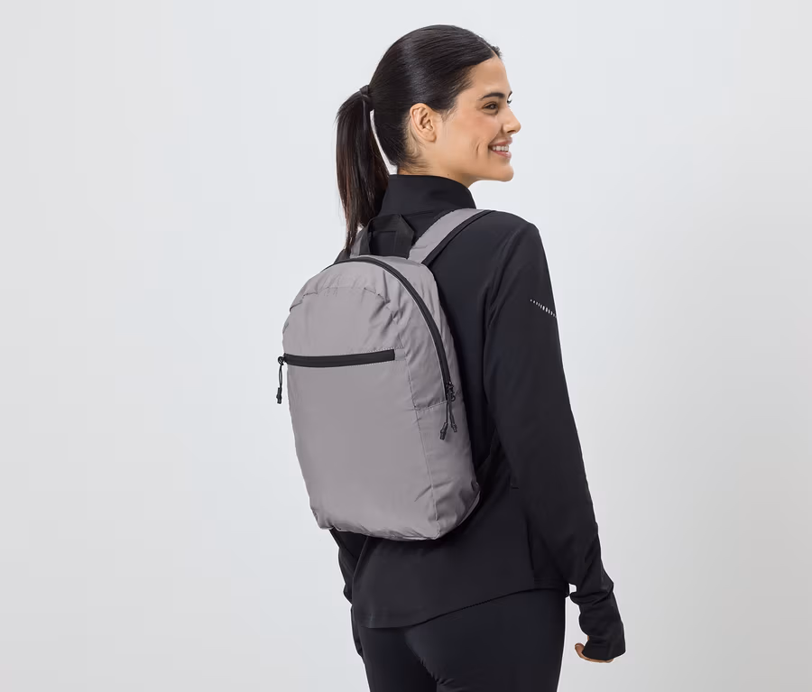 Une femme avec une queue de cheval porte un sac à dos gris et des vêtements de sport noirs, vue de côté.