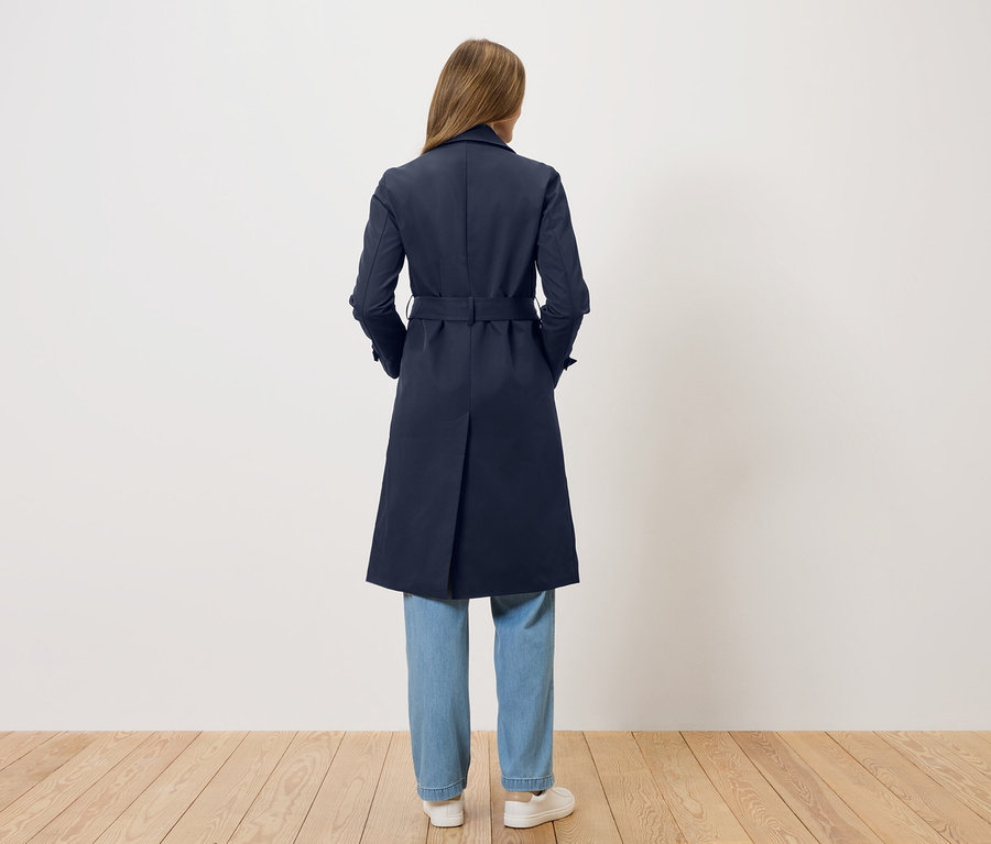 Une femme, dos à la caméra, porte un trench-coat bleu marine, un jean et des chaussures blanches.