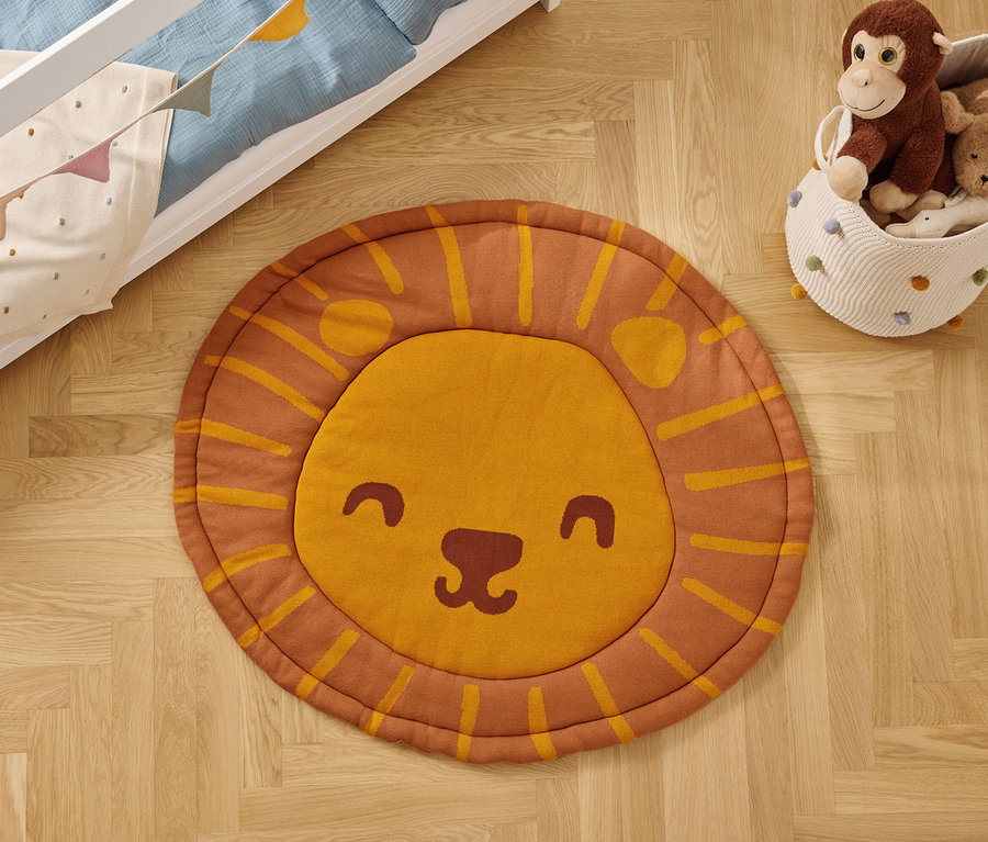 Sur un plancher en bois, on trouve un tapis rond avec une tête de lion, à côté d'un panier à jouets et d'un lit d'enfant.