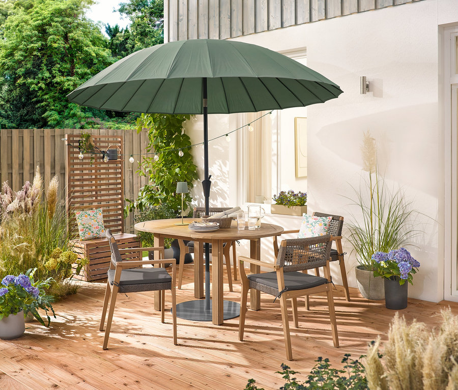 Une table ronde en bois avec quatre chaises de jardin empilables avec un tressage textile se trouve sous un parasol vert. Une guirlande lumineuse solaire »Glühbirnen« est suspendue au-dessus de la table, sur laquelle se trouve une lampe de table d'extérieur LED à batterie, sauge. Deux coussins d'extérieur à motifs floraux sont posés sur une caisse en bois.
