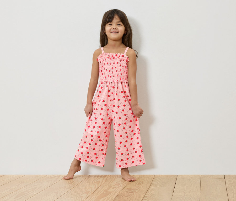 Une jeune fille se tient pieds nus devant un mur blanc, portant une combinaison rose à motifs de fraises.