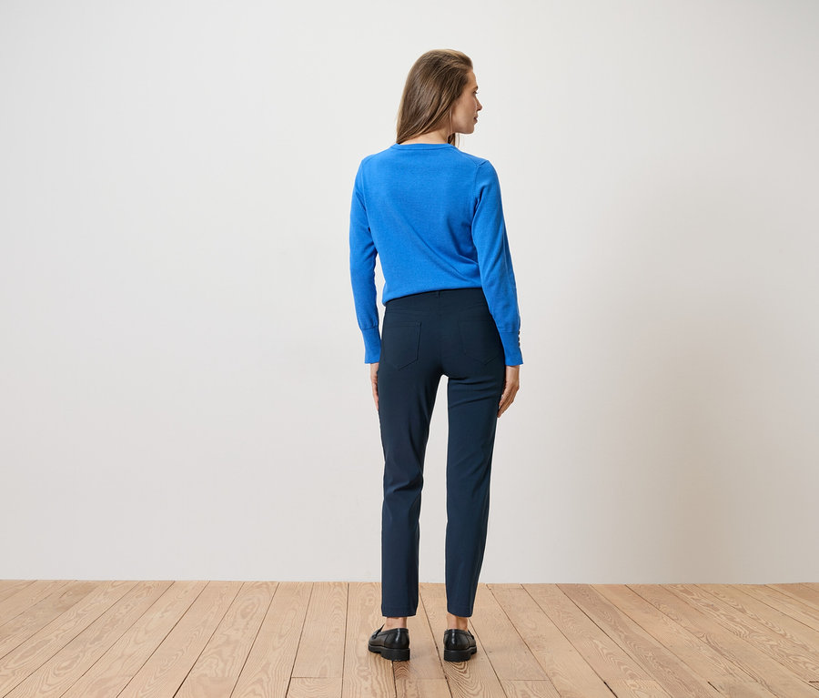 Une femme vue de dos, portant un pull bleu, un pantalon bleu et des chaussures noires, se tient sur un plancher en bois devant un mur blanc.