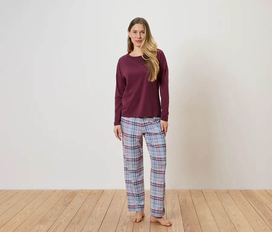 Une femme se tient pieds nus devant un mur blanc, portant un pull bordeaux et un pantalon de pyjama à carreaux bleus.