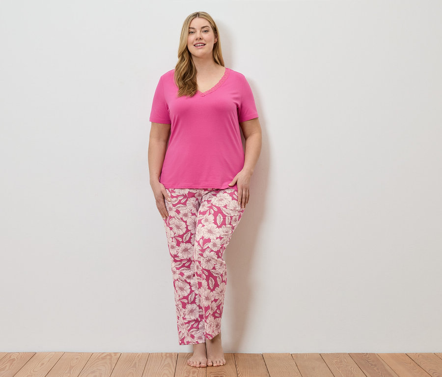Une femme se tient pieds nus devant un mur blanc, portant un pyjama rose à motifs floraux.