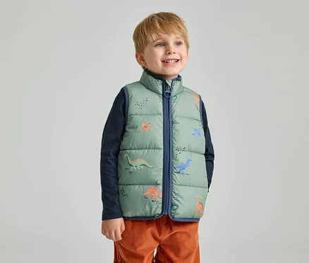 Un garçon souriant aux cheveux blonds courts porte un gilet matelassé vert sans manches avec des motifs de dinosaures.