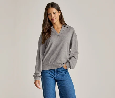 Une femme aux cheveux bruns porte un pull gris à col et un jean bleu, une main dans la poche.