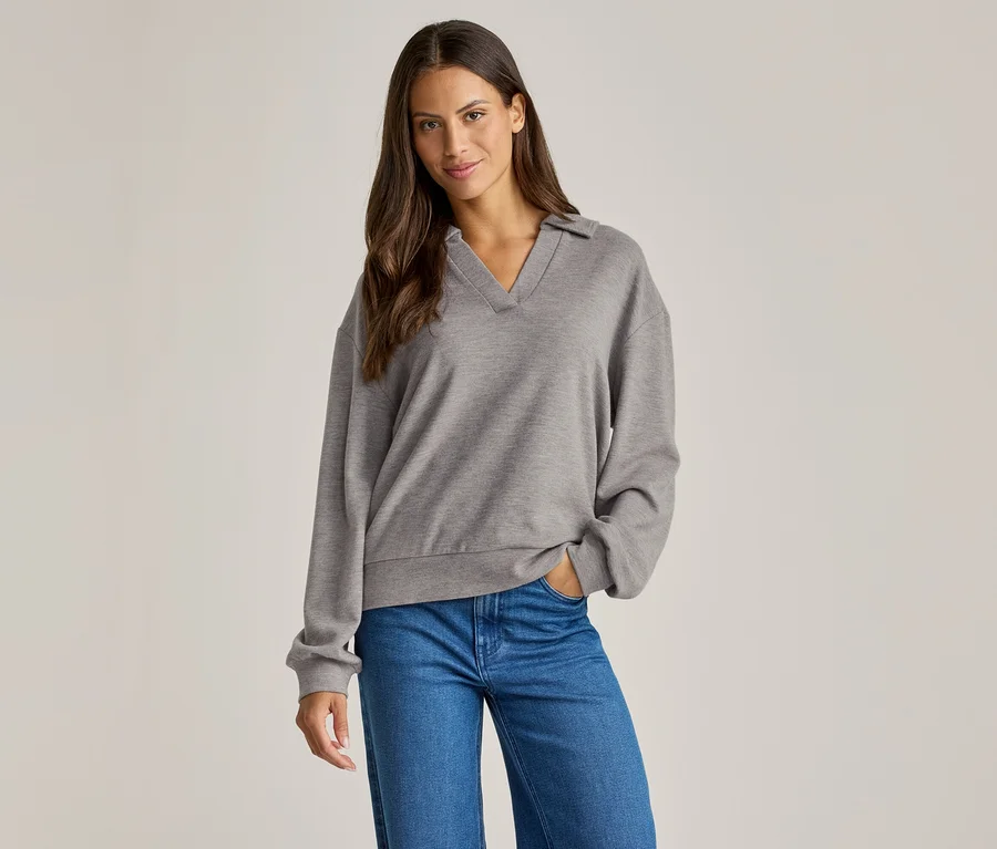 Une femme aux cheveux bruns porte un pull gris à col et un jean bleu, une main dans la poche.