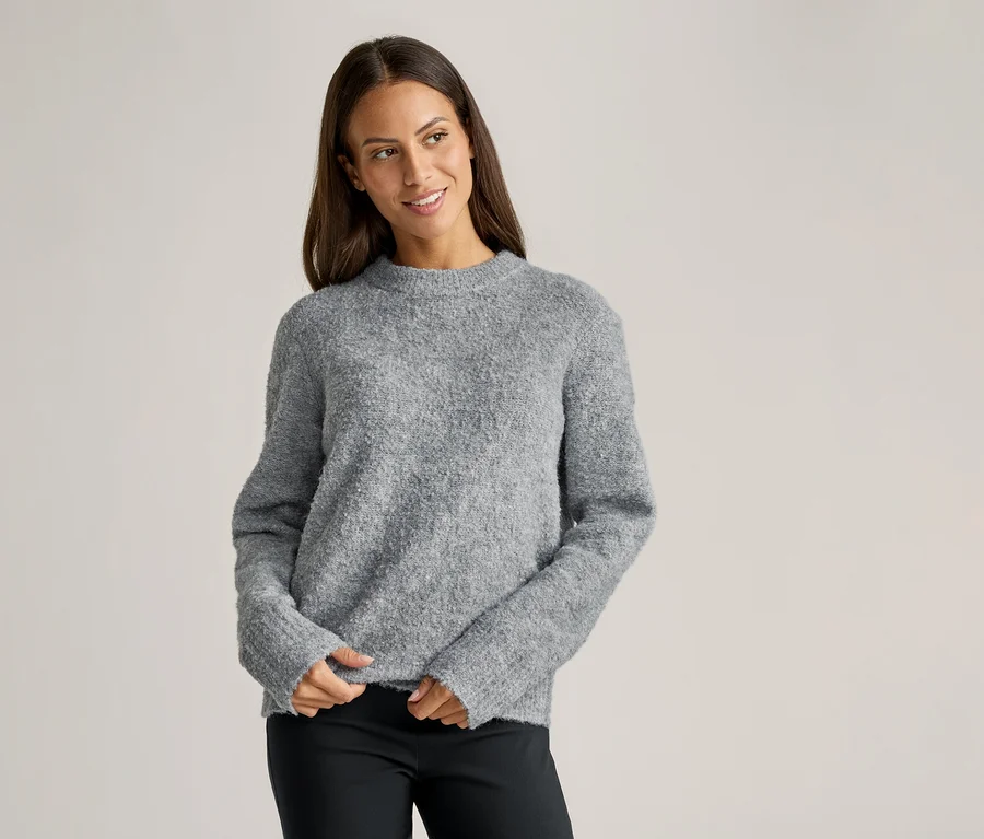 Une femme aux cheveux bruns porte un pull gris et un pantalon noir sur un fond clair.