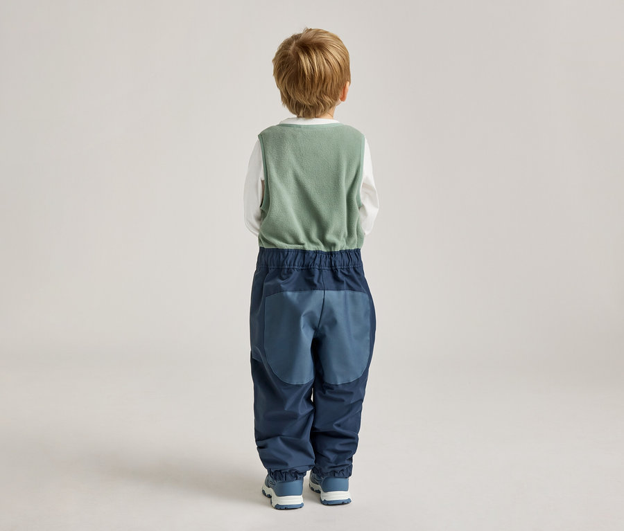 Vue arrière d'un enfant blond portant un gilet vert, un t-shirt blanc à manches longues et un pantalon bleu à blocs de couleurs.