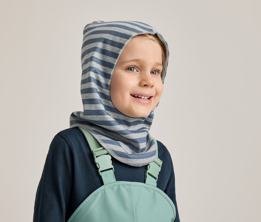 Un garçon souriant portant une cagoule rayée bleu et gris, un t-shirt à manches longues bleu marine et une salopette vert menthe sur fond gris clair.