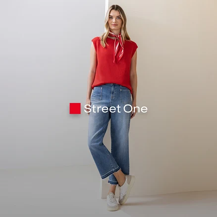 Une femme portant un haut rouge et un jean, avec un foulard rouge et blanc autour du cou, pose devant un fond clair.