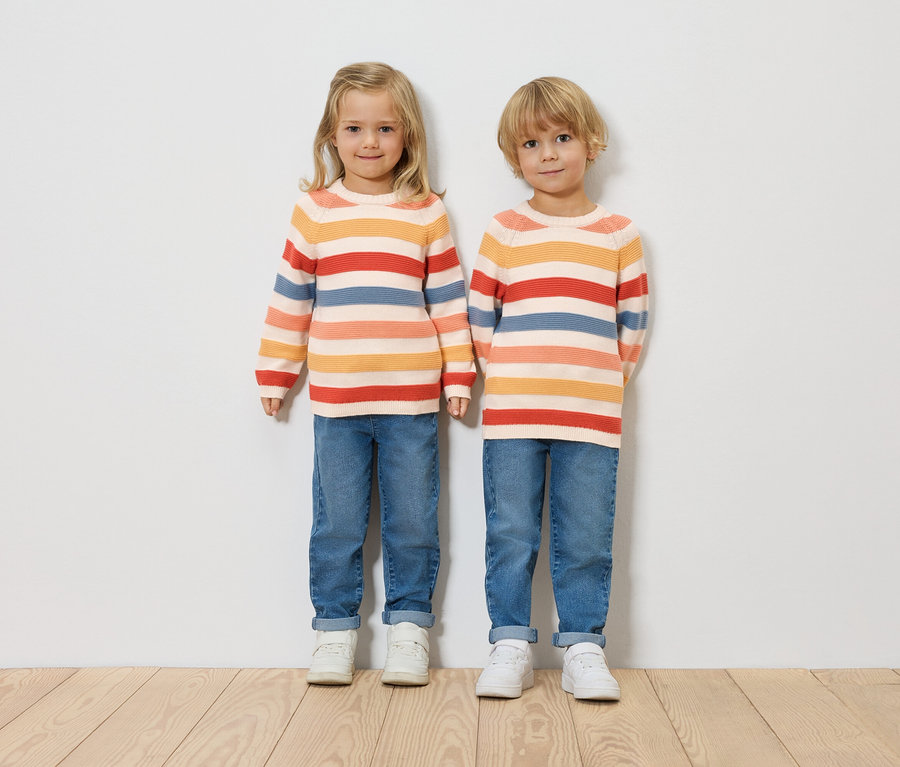 Deux enfants se tiennent côte à côte devant un mur blanc. Ils portent des pulls rayés, des jeans et des baskets blanches.