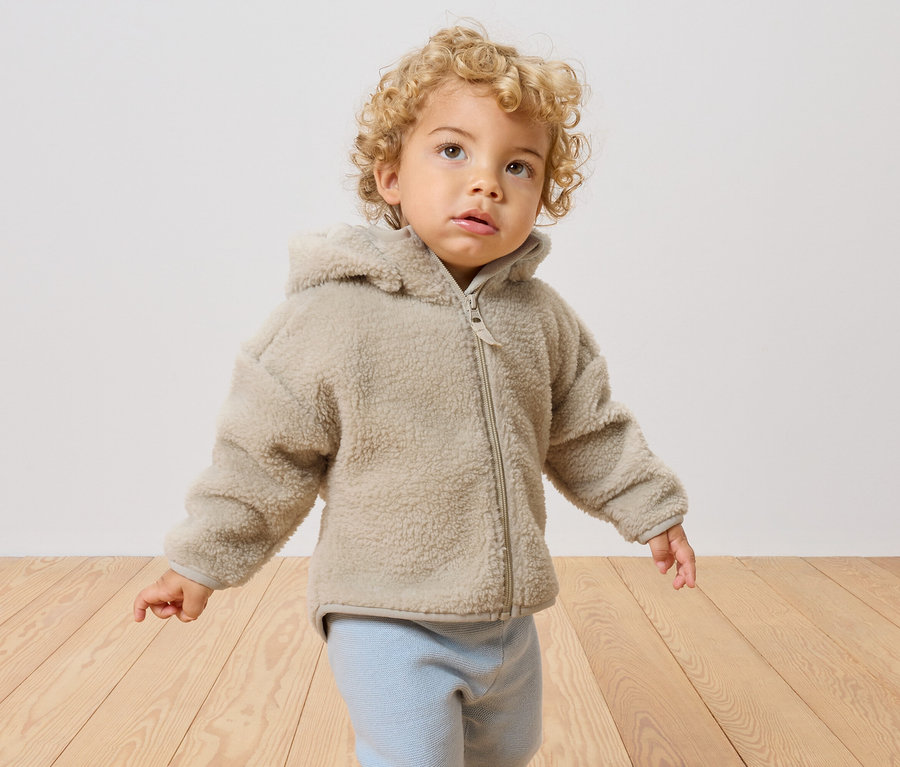 Un jeune enfant aux cheveux blonds bouclés porte une veste en teddy beige avec capuche et un pantalon bleu clair.