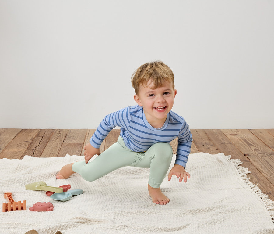 Un garçon souriant en haut rayé bleu et leggings vert clair s'étire sur une couverture blanche à côté de jouets.