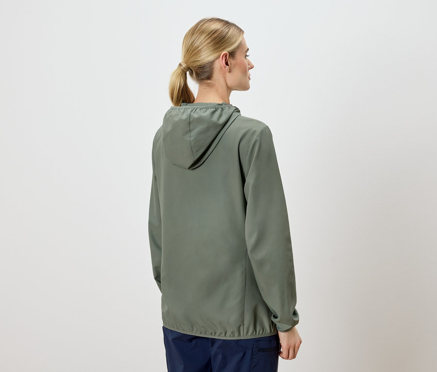 Vue arrière d'une femme blonde avec une queue de cheval portant une veste à capuche vert olive et un pantalon bleu marine sur un fond clair.