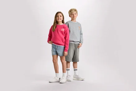 Une fille portant un pull rose et un short en jean se tient à côté d'un garçon portant un pull gris et un short.