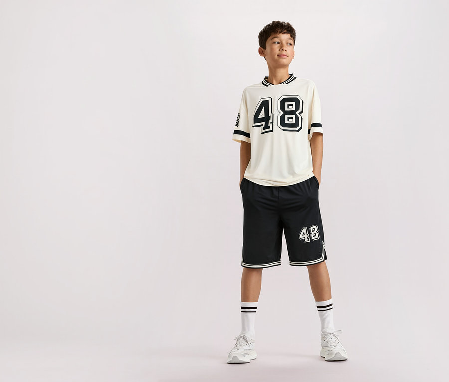 Un adolescent se tient debout, les mains dans les poches, vêtu d'un t-shirt beige avec le numéro 48, d'un short noir avec le numéro 48, de chaussettes blanches rayées et de baskets blanches sur un fond uni.