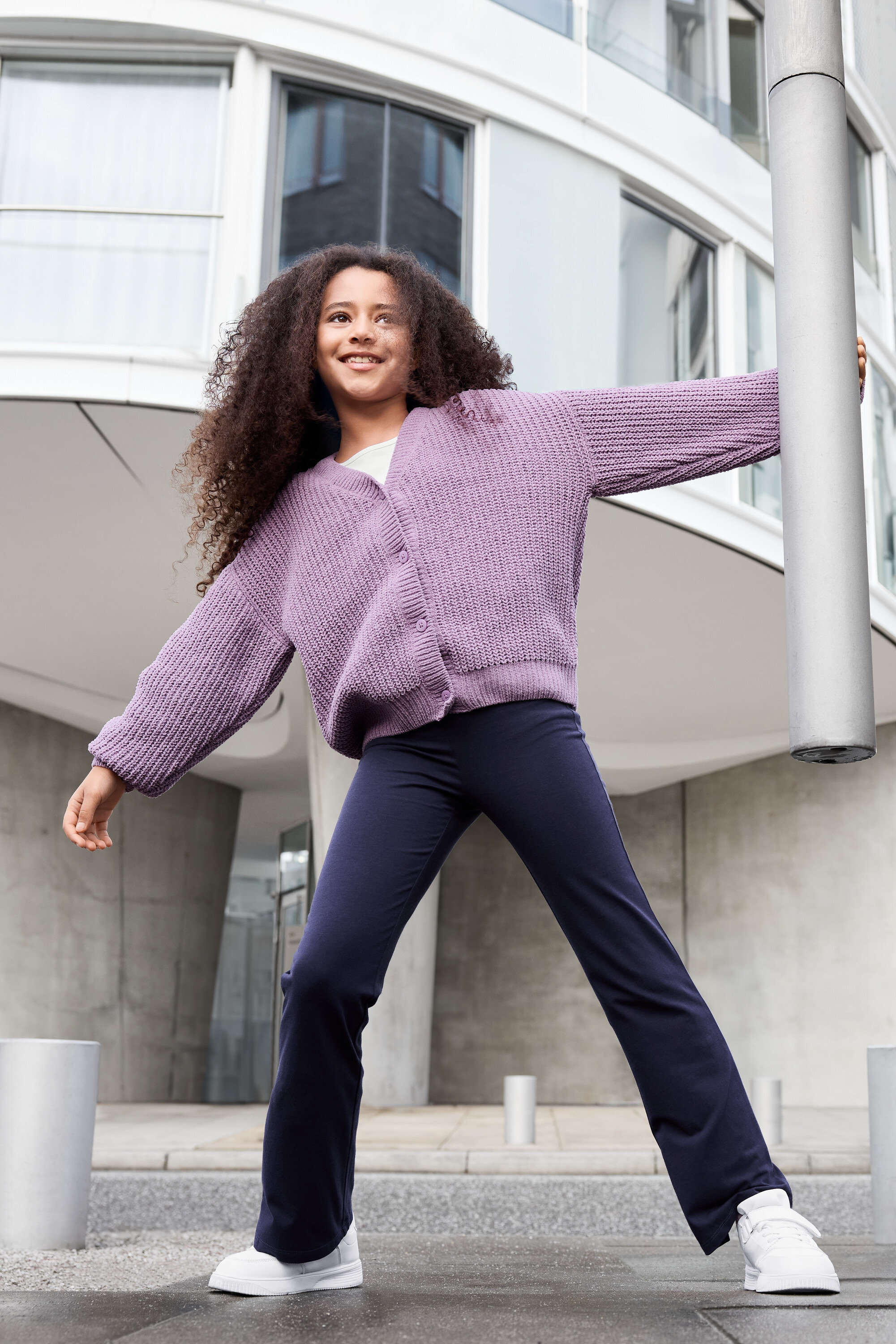 Une fille aux cheveux bouclés, portant un cardigan lilas, un pantalon bleu et des baskets blanches, pose devant un immeuble.