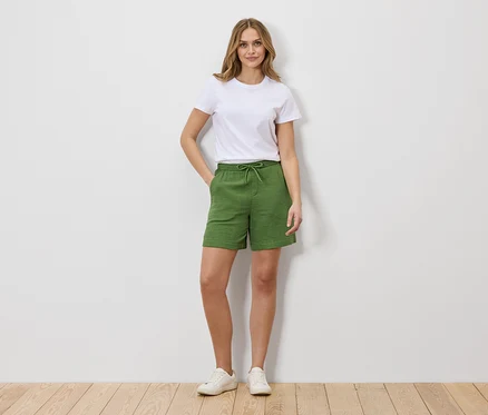 Une femme en t-shirt blanc et short vert à cordonnet se tient devant un mur blanc.