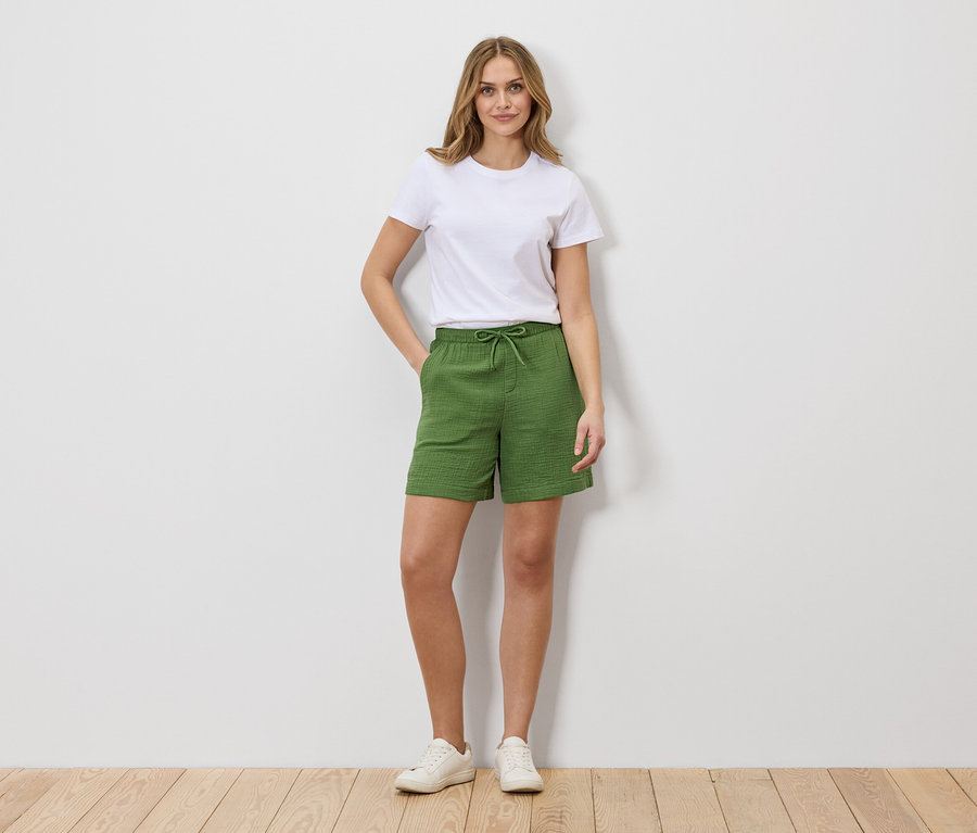 Une femme en t-shirt blanc et short vert à cordonnet se tient devant un mur blanc.