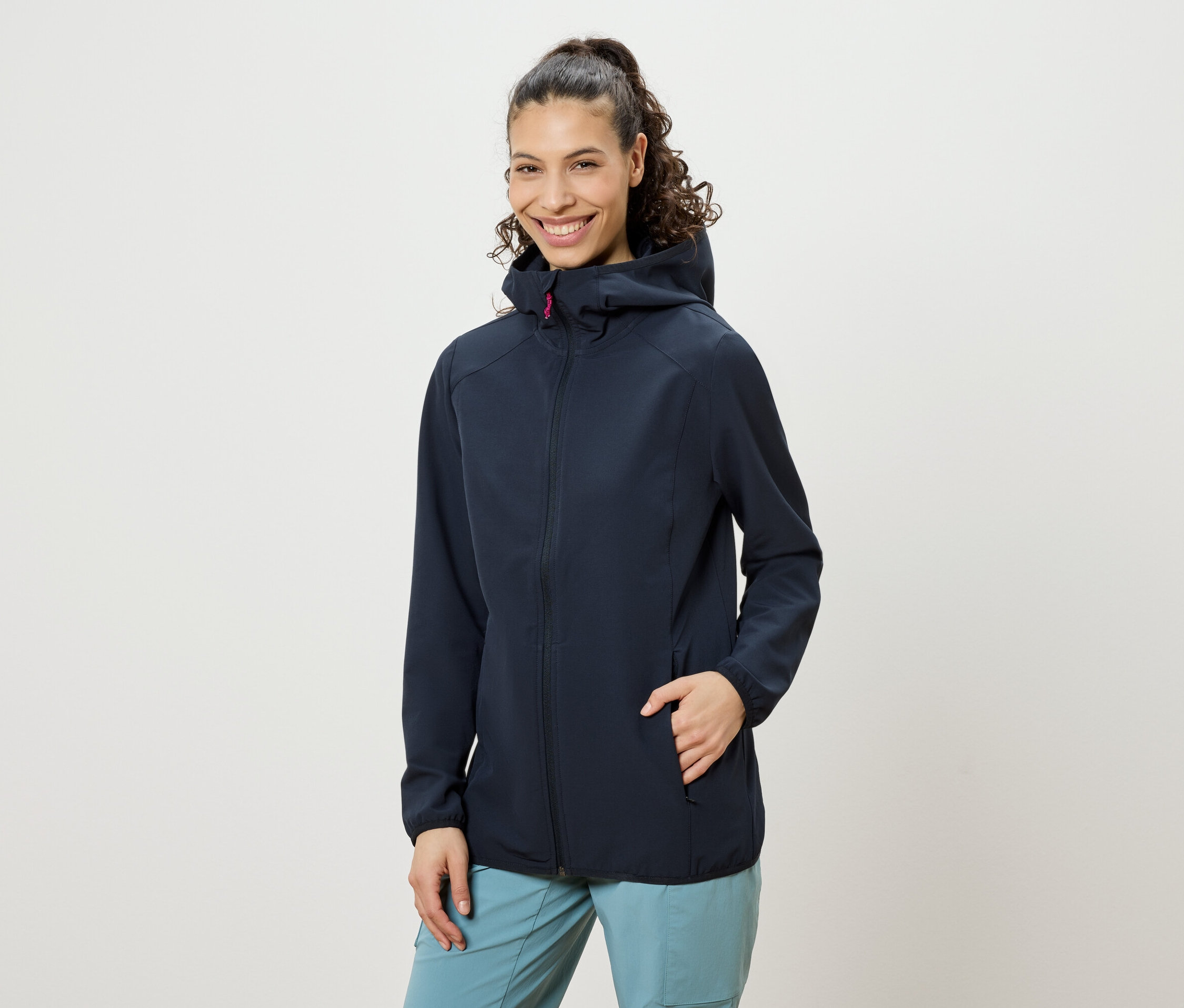 Une femme souriante aux cheveux bouclés remontés en queue de cheval, portant une veste à capuche bleu marine et un pantalon bleu, avec une main dans la poche de sa veste.