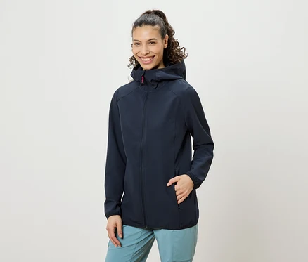 Une femme souriante aux cheveux bouclés remontés en queue de cheval, portant une veste à capuche bleu marine et un pantalon bleu, avec une main dans la poche de sa veste.