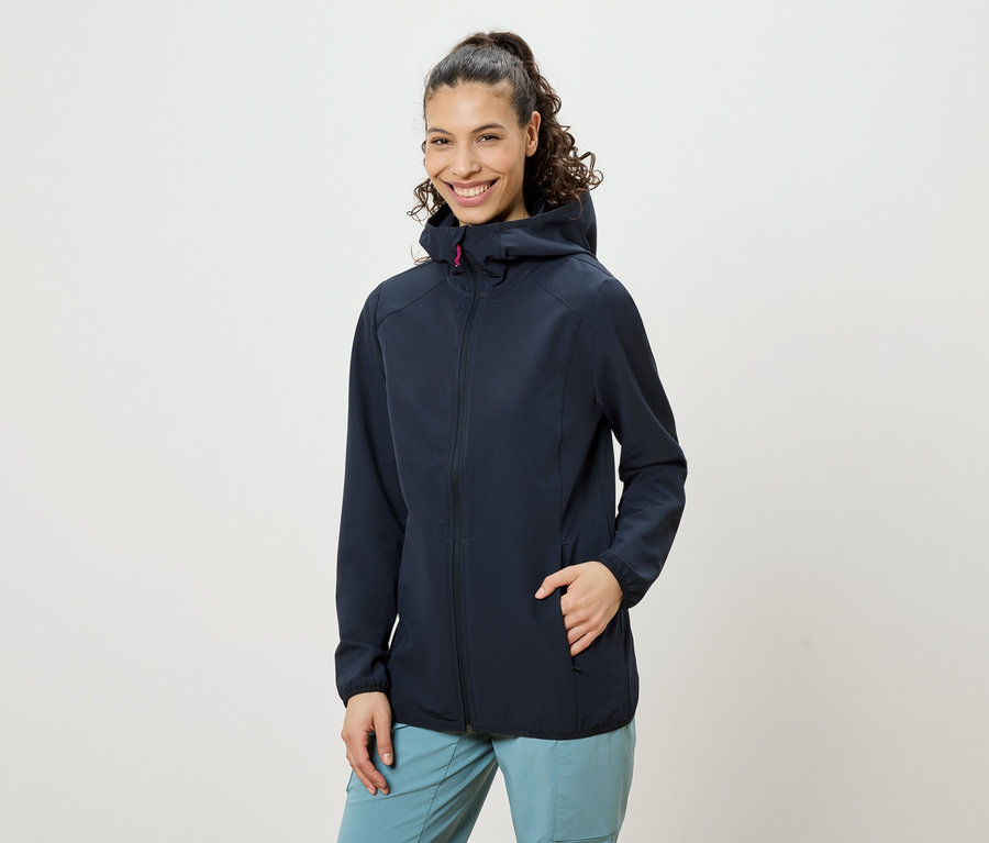 Une femme souriante aux cheveux bouclés remontés en queue de cheval, portant une veste à capuche bleu marine et un pantalon bleu, avec une main dans la poche de sa veste.