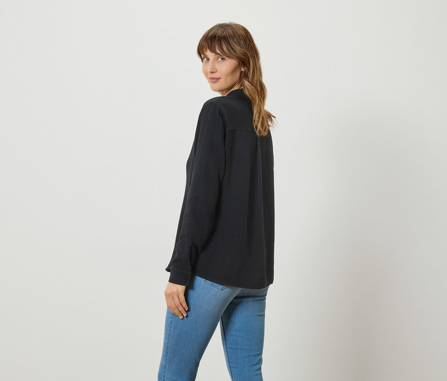 Femme aux longs cheveux ondulés, portant une blouse longue et foncée et un jean, regardant par-dessus son épaule vers la caméra.