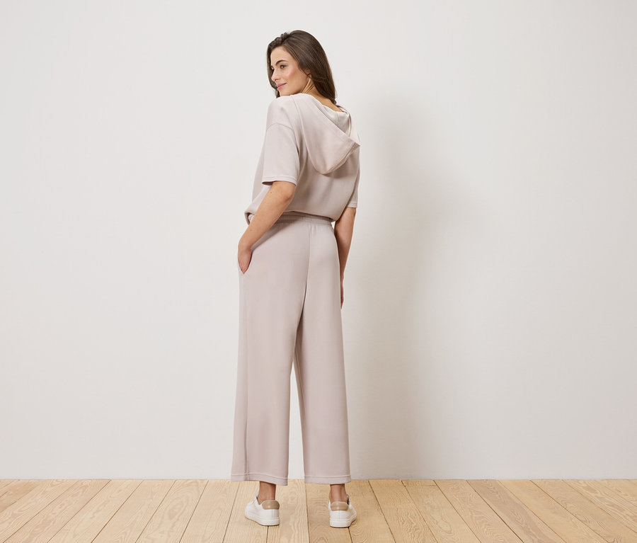 Une femme aux cheveux bruns, photographiée de dos, porte un haut à capuche marron clair et un pantalon large.