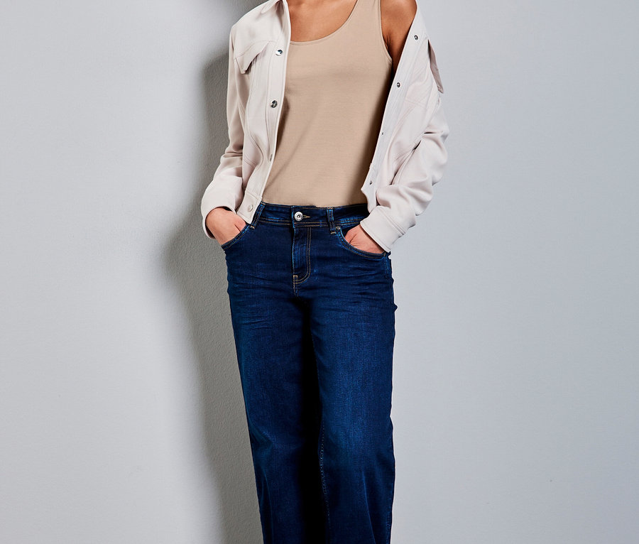 Femme portant un haut classique Street One beige, un jean large bleu foncé et une veste claire sur les épaules, les mains dans les poches.