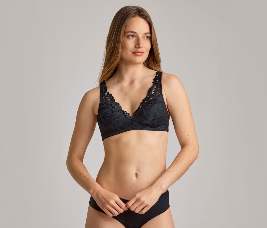 Une femme aux longs cheveux bruns porte un soutien-gorge en dentelle noire et une culotte noire sur un fond gris.
