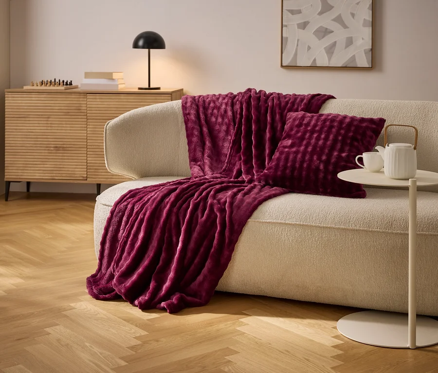 Un plaid et un coussin bordeaux sont posés sur un canapé crème. Une table d'appoint avec une théière et une tasse se trouve à côté.