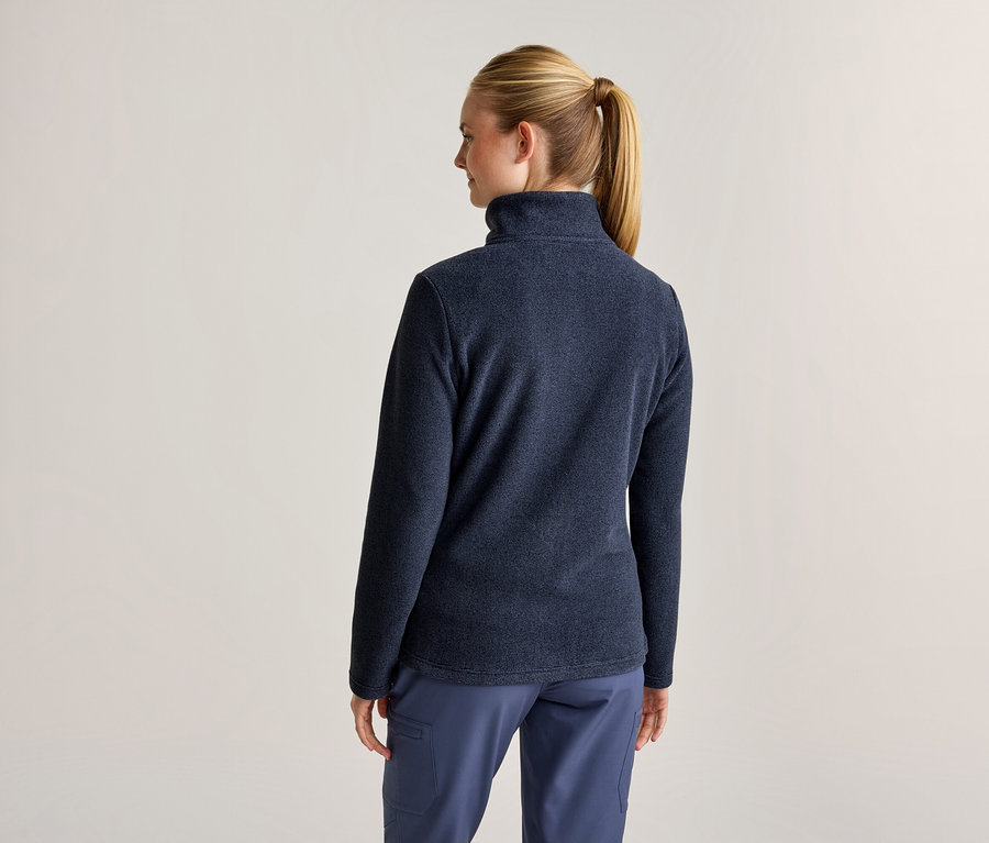 Une femme vue de dos, portant une veste polaire bleu foncé et un pantalon bleu. Ses cheveux blonds sont attachés en queue de cheval.