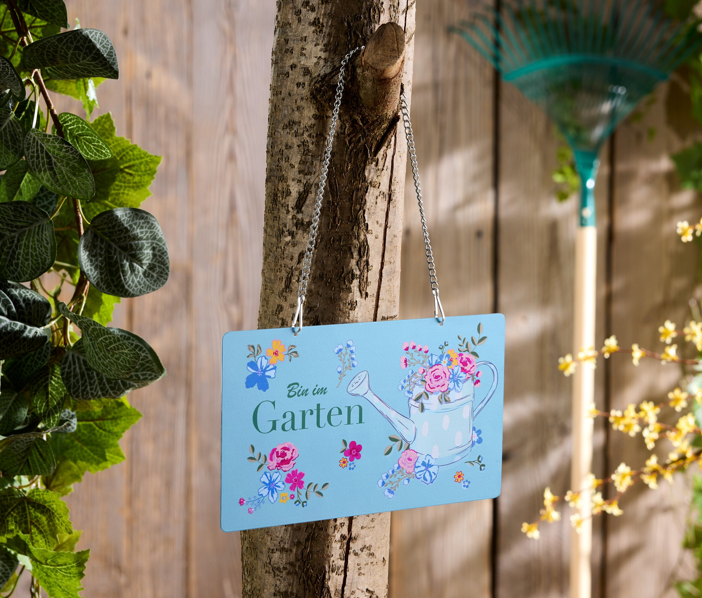 Un panneau bleu avec l'inscription "Bin im Garten" est accroché à un arbre, un râteau en arrière-plan.